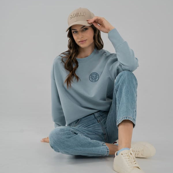 Woman wearing a light blue SOMI sweatshirt, jeans, and beige cap on a gray background