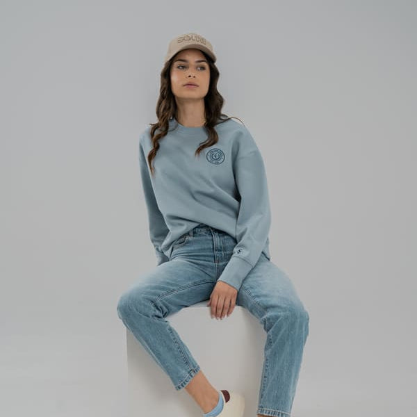 Woman sitting on a white cube wearing a light blue SOMI sweatshirt, jeans, and beige cap against a plain background