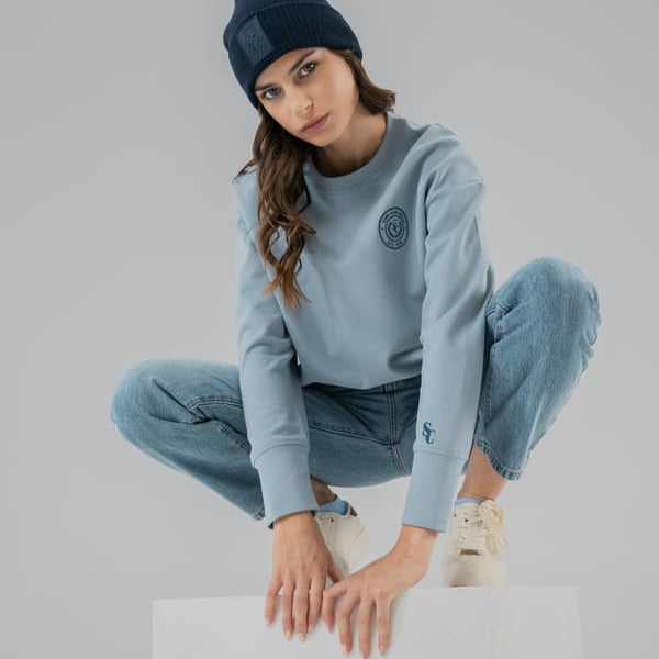 Woman wearing a light blue SOMI sweatshirt and pants with a dark beanie on a gray background