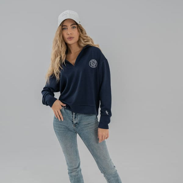 Woman wearing a navy blue SOMI sweatshirt, light blue jeans, and white sneakers on a gray background