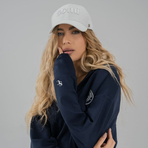Woman wearing a navy blue SOMI sweatshirt and white cap with a logo on a gray background