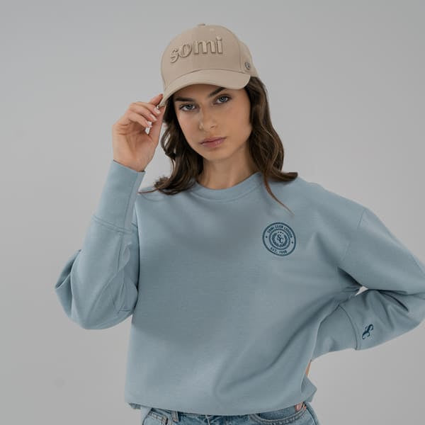 Woman wearing a light blue SOMI sweatshirt and jeans, holding a beige cap with text.
