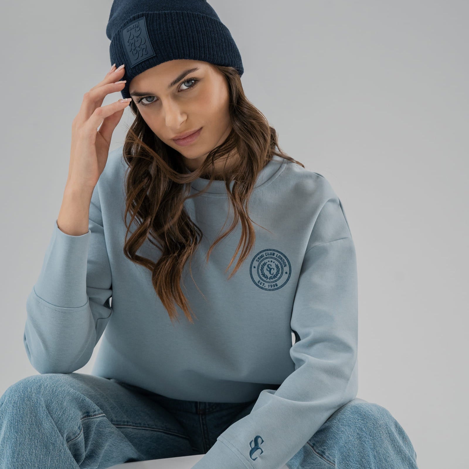 Woman wearing a light blue SOMI hoodie with a logo, sitting on a white chair against a gray background